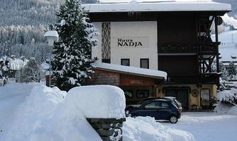 A charming, snow-covered house named "Haus Nadja". Surrounded by snow-covered trees and mountains.
