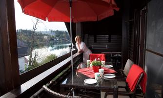 A cozy balcony with a beautiful view of the water. A woman is leaning on the balustrade and the table is set with red dishes.