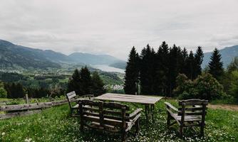 A viewpoint with wooden chairs and a table on a meadow. In the background, mountains and a lake stretch out under a cloudy sky.