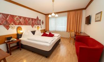 A cozy hotel room with a double bed and red accents. There is a desk, a TV, and plenty of daylight coming through the window.