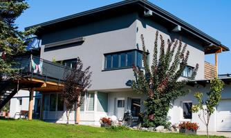 A modern house with a gray facade and large windows. It is situated in a green environment with trees and flowers.
