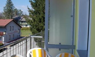 A balcony with a round table and four chairs in yellow and white. In the background, houses and trees are visible.