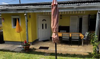 A yellow house with a terrace and a table with chairs. In the foreground, there are umbrellas and flowers in a pot.