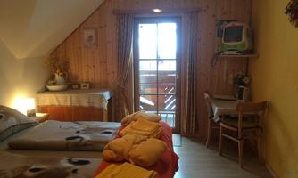 A cozy bedroom with wooden paneling and a window that leads to a balcony. Colorful towels are laid on the bed and a small seating area with a table and chairs is available.