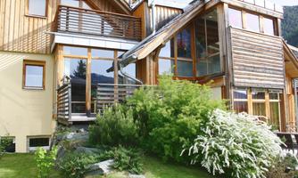 A modern wooden house with large windows and a terrace. In the foreground, green plants and shrubs can be seen.