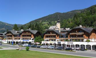 A picturesque village with traditional, wood-clad buildings. In the background, green hills extend and a blue sky.