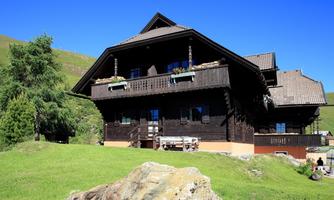 A traditional wooden house with a beautiful balcony. It stands in a green, hilly landscape under clear, blue skies.