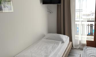 A simple hotel room with a single bed and a television on the wall. There is a balcony with curtains and plenty of daylight.