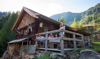 A cozy mountain cabin surrounded by green forests and mountains. Several people are sitting on the porch, enjoying the view.