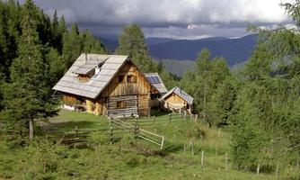 A rustic cabin surrounded by trees and meadows in the mountains. The sky is partly cloudy and the landscape is green and picturesque.