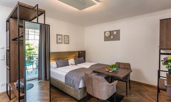 A modern hotel room with a double bed and a small table. The decor is simple and elegant, with wooden flooring and a window that lets in a lot of light.