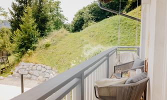 A balcony with comfortable chairs and a view of a green meadow. In the background, there are large trees and the sky is clear.