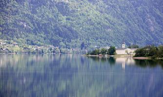 A tranquil sea landscape with reflecting water and gentle hills. In the background, a charming building can be seen among the trees.