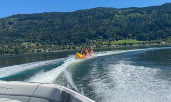 A group of people is riding on a banana boat across a clear lake. In the background, green mountains and an idyllic landscape can be seen.