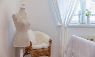 An elegant dress form stands next to a wicker armchair in a bright room. White curtains and floral decorations add a cozy atmosphere.