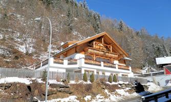 A beautiful house in alpine style, surrounded by snow-covered mountains. The clear sky and the trees give the scene a peaceful atmosphere.
