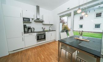 A modern kitchen with white cabinets and appliances. A dining table with chairs stands by the window, overlooking a green outdoor area.