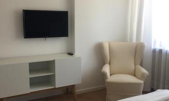 A bright bedroom with a cozy armchair and a television on the wall. The room has large windows that let in plenty of daylight.