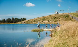 A group of people is hiking along a calm lake. In the background, gentle hills and a blue sky with a few clouds can be seen.