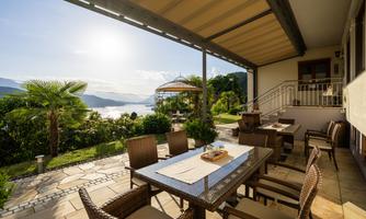 A cozy terrace with a table and chairs offers a view of the lake and the surrounding landscape. The sun is shining and there is a relaxed atmosphere.