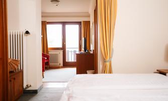 A cozy hotel room with a view of a balcony. The windows are framed by light curtains and the room is simply furnished.