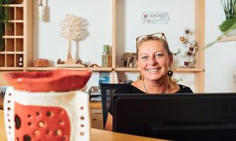 A friendly woman is sitting at a desk with a computer. In the background, decorative elements and plants are visible.