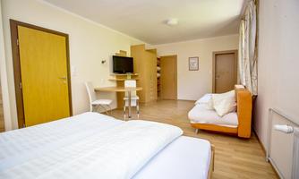 A modern room with a double bed, a table, and a television. It has bright walls and a light wooden floor.