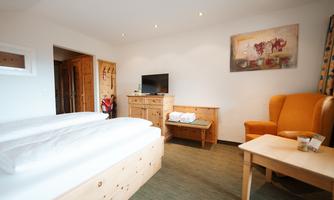 A bright hotel room with two beds, a television, and a cozy armchair. There is a wooden table and a decorative wall with a picture.