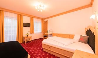 A cozy room with wooden decor, a large bed, and a television. Bright curtains let in plenty of daylight.