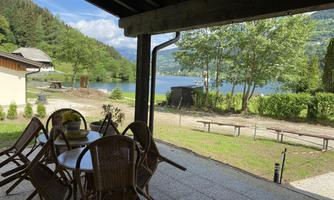 A cozy terrace with a round table and chairs, overlooking a tranquil lake and green trees in the surroundings. The sky is clear and nature is harmonious.