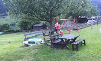 A well-maintained garden with a swing, a picnic table, and a large tree. In the background, huts and gently rolling hills can be seen.