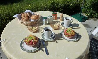A set table outside with fresh bread, two cups of coffee, and fruit salad. The table is surrounded by green plants.