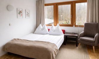 A cozy bedroom with a double bed and two pillows.  
Large windows offer a view outside and let in plenty of daylight.