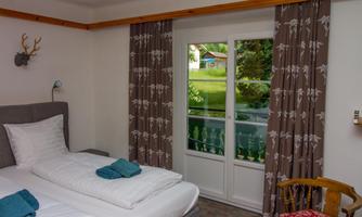 A cozy room with two single beds, a small table, and a beautiful view of the garden through large windows. The walls are painted in neutral colors, complemented by decorative curtains.