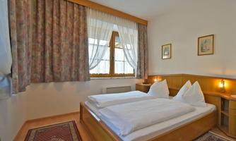 A cozy bedroom with two beds, padded pillows, and a window with curtains. Pictures hang on the walls, and a rug lies on the floor.