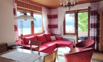 A cozy living room with a red sofa and a red armchair. Large windows offer a view of the lake and let in plenty of light.