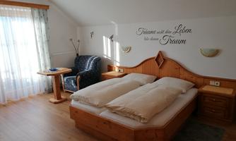 A cozy bedroom with a double bed, wooden furniture, and a reading chair. Bright curtains create a friendly atmosphere.