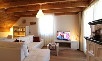 A cozy living room with a white couch and wooden beam ceiling. A television displays a colorful program, while large windows let in plenty of daylight.