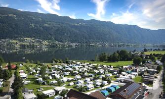 A picturesque campsite by the lake, surrounded by green forests and mountains. The sky is clear and the landscape is calm and inviting.