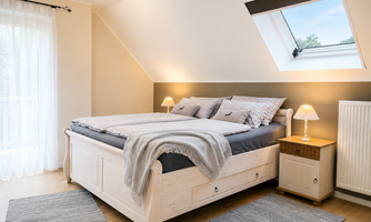 A cozy bedroom with a large bed and soft color tones. The windows let in a lot of light and create an inviting atmosphere.