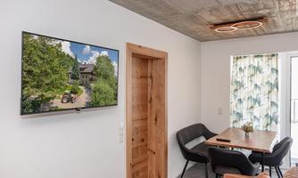 A modern living room with a flat-screen TV on the wall. The table is equipped with black chairs and a large window with a view of nature.