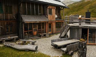 A rustic wooden house in a mountainous setting. Outside, there is a terrace with wooden furniture and a stone area.