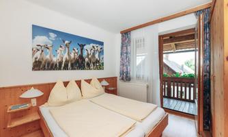 A cozy bedroom with a large bed and wooden paneling. The wall is decorated with a picture of cows, and the window provides access to a balcony.