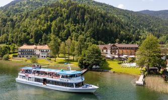 A ship sails on a calm lake, surrounded by green forests and hills. In the background, charming buildings and a sunny landscape can be seen.