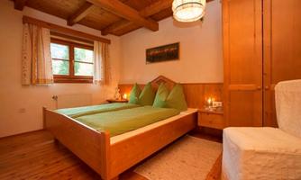 A cozy bedroom with a wooden bed and green cushions. The room features wood paneling and large windows that let in light.