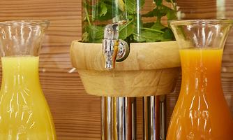 A decorative drink dispenser with fresh herbs and flowers. Next to it are two pitchers with yellow and orange juice.