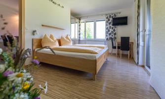 A bright, modern bedroom with a double bed and large windows. Next to the bed is a desk with a television in the corner.