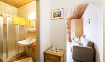 A bright bathroom with a shower, sink, and towels in warm colors. In the background, a cozy living area with a sofa and decorations can be seen.