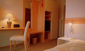 A simple room with wooden furniture, a desk, and a comfortable chair. There is a table lamp and a small fridge nearby.