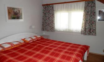 A cozy bedroom with a large bed and checkered bedding. The room is bright and friendly, with floral pictures on the walls and colorful curtains.